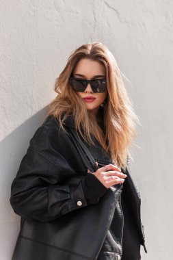 Moda gözlüklü, şık, siyah deri ceketli, şık, genç, hippi bir kız. Güneşli bir bahar gününde, gri bir duvarın yanında, sokakta büyük siyah bir çanta.