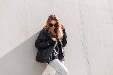 Moda modası geçmiş havalı hippi kız modeli rock tarzı deri ceketli, kot pantolonlu ve modern güneş gözlüklü şık çanta güneşli bir günde gri bir duvarın yanında duruyor.