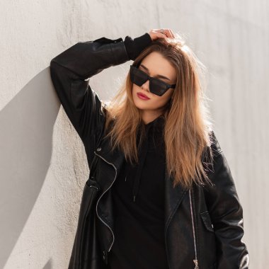 Moda modası geçmiş, havalı güneş gözlüklü, deri ceketli ve kapüşonlu hippi bir kadın sokakta gri bir duvarın yanında duruyor. Kadın bahar stili, moda ve güzellik