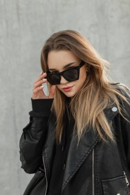 Moda kıyafetleri içinde, deri ceketli ve kapüşonlu güzel bir kızın, gri bir duvarın yanında güneş gözlüğünü ayarlayan şık bir portresi.