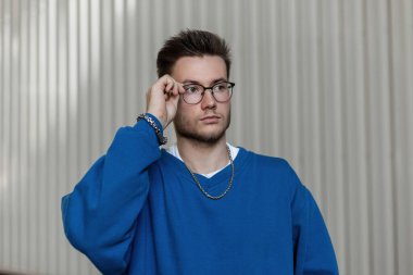 Modaya uygun mavi kazaklı şık genç hippi sokakta metalik gri bir arka planda gözlük takıyor.