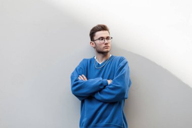 Moda gözlüklü, saç stili ve modaya uygun mavi bir sweatshirt giyen hippi stili genç adam sokakta beyaz duvarın yanında el ele duruyor ve bakıyor.