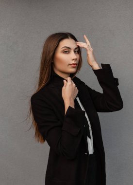 Siyah ceketli, beyaz gömlekli ve kravatlı şık siyah iş elbiseli güzel bir kız gri bir duvarın yanında duruyor.