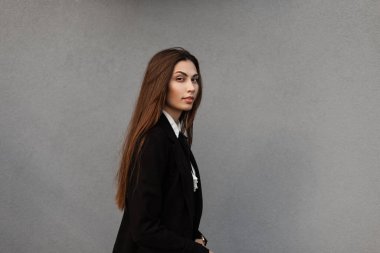 Şık siyah elbiseli, gömlekli, ceketli ve kravatlı güzel bir kız model. Gri duvarın yanında, şehirde yürüyor.