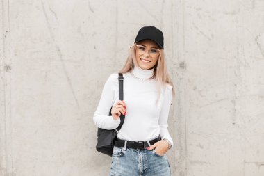 Şık, sıradan şapkalı, şık bir kadın. Gözlük, süveter, kot pantolon ve siyah sırt çantası gri beton arka planda