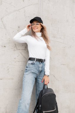 Süslü beyaz bir süveter, kot pantolon ve kepiyle gülümseyen güzel bir genç kız sırt çantası tutuyor ve beton bir duvarın yanında duruyor.