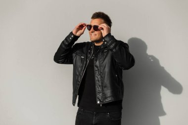 Gülümseyen, havalı siyah elbiseli, deri ceketli havalı vahşi hippi şehirde gri bir arka planda siyah gözlük takıyor.