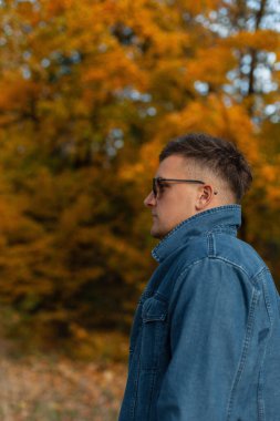 Sonbahar parkında yürüyen kot ceketli, saç stili ve güneş gözlüklü yakışıklı bir adamın modaya uygun sonbahar portresi.