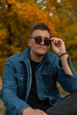 Moda giysileri içinde şık genç adam modeli sonbahar parkında oturur ve klasik güneş gözlüğü takar. Sonbahar erkeği tarzı kıyafetler.
