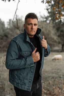 Şık mavi kot ceketli, siyah tişörtlü ve kotlu genç adam kırsal alanda duruyor.