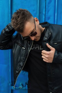 Havalı, modaya uygun, siyah kıyafetli, siyah deri ceketli ve gözlüklü genç hippi mavi klasik metal bir duvarın yanında duruyor.