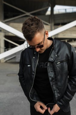Güneş gözlüklü şık siyah kıyafetli genç yakışıklı adam deri ceketinin fermuarını çekip kasabada dolaşıyor.