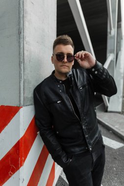 Siyah elbiseli, siyah ceketli, güneş gözlüklü yakışıklı hippi sokakta duruyor.