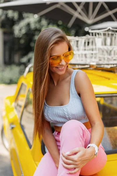 Funny beautiful young hipster girl with a positive summer vibe in fashionable colored clothes with glasses, knitted top and pink denim sits and enjoys herself on a yellow retro car