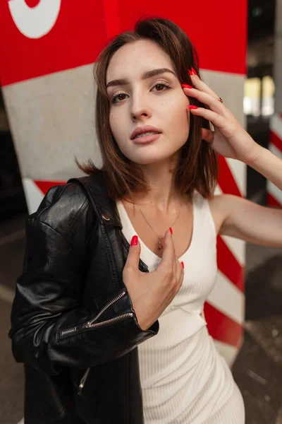 穿着皮夹克 身穿白色休闲装 长相俊俏的时尚时髦女性模特出现在混凝土红墙旁的大街上 — 图库照片