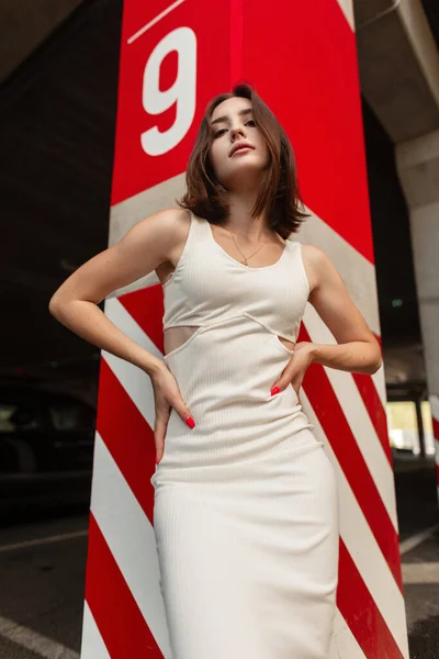 白いドレスの髪型をしたかなりセクシーな女性モデルが立っており 通りの赤白のコンクリート列の近くにポーズをとっています 都会的なスタイル ファッション 美しさ — ストック写真