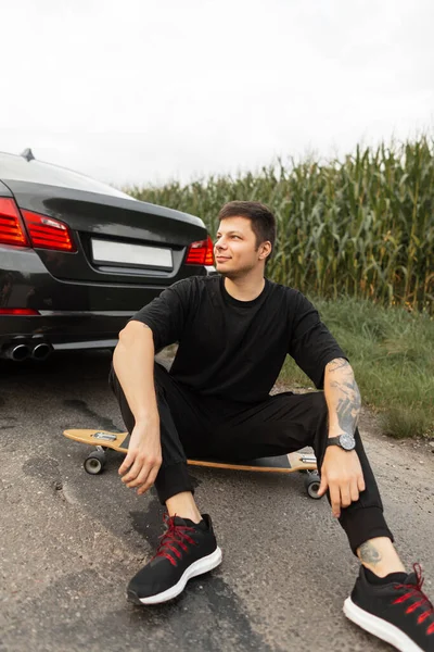 Hipster man in fashion black clothes with sneakers sits on longboard near a black car and rest