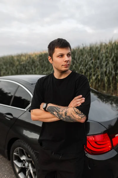 Stylish handsome hipster guy in a black T-shirt with a tattoo on arm standing by car on the road outside the city