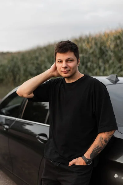 Hipster man with tattoo on hand in black fashion t-shirt and jeans stands near a car outdoors