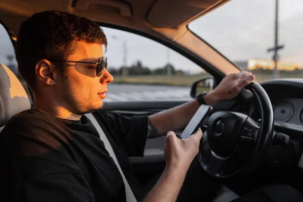 一个戴墨镜 身穿黑色T恤的英俊商人正在使用智能手机 并在日落时开着汽车 — 图库照片
