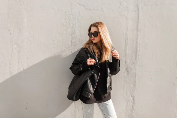 时尚的年轻貌美的女人站在街上灰蒙蒙的墙上 戴着太阳镜 穿着凉爽的皮夹克 帽衫和牛仔裤 头戴皮制黑色包 — 图库照片