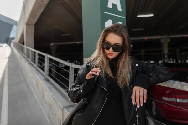 Fashionable young beautiful woman model hipster  in leather black rock jacket and black hoodie with handbag walks on the city. Urban female style look clothes