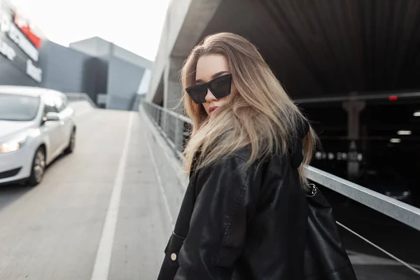 Premium Photo  Hipster beautiful girl with fashion sunglasses in trendy  leather jacket and black hoodie poses on the street near a parking lot  urban casual clothes fashion and female style