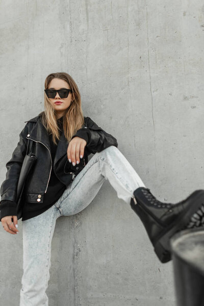 Stylish beautiful young hipster woman with sunglasses in a fashion casual outfit with a leather jacket, hoodie, jeans and boots with a bag posing on the street near a gray wall