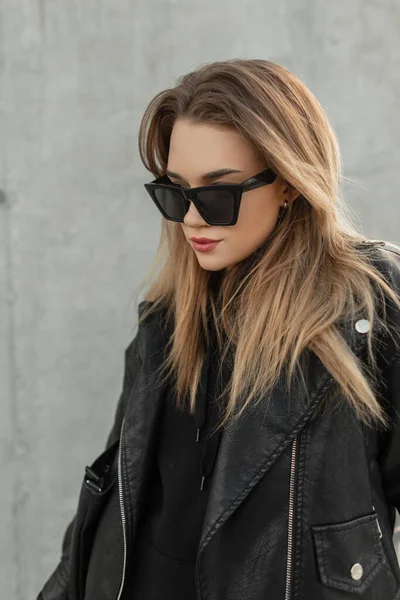 Fashionable young woman model hipster with hairstyle and stylish sunglasses in trendy black rock leather jacket and black hoodie stands near a gray wall
