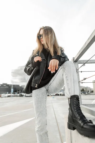 Cool rock fashionable girl with trendy fashion leather black jacket, jeans and black stylish shoes with leather bag poses in the city. Urban fashion and beauty
