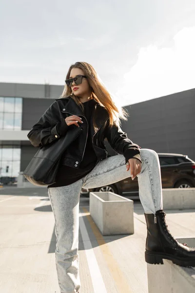 beautiful fashionable girl in black rock style with a black leather jacket, jeans, sunglasses and stylish boots with a bag poses in the city. Women\'s urban clothing style