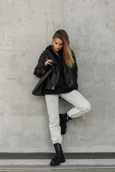 Cool fashionable woman hipster with hairstyle in trendy spring outfit with leather jacket, jeans and leather black boots stands and poses near a gray wall on the street