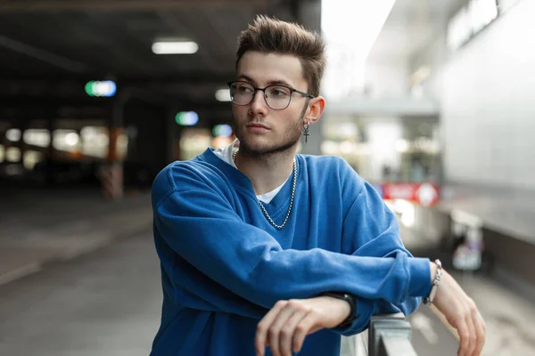 Hipster young man with hairstyle and glasses in blue fashionable sweatshirt stands in the city. Urban male style look clothes