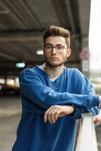 Handsome fashionable business man hipster with hairstyle and glasses in stylish blue sweatshirt stands in the city