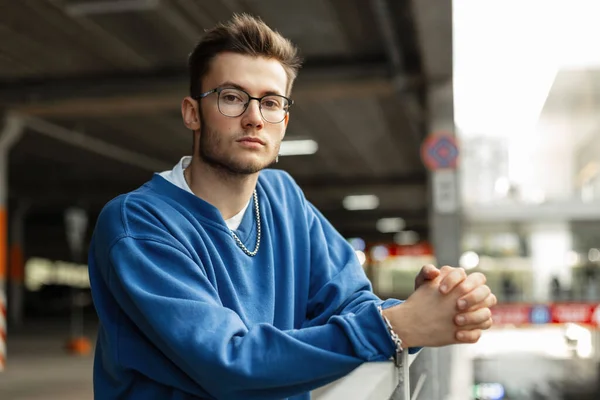 Fashion handsome man with hairstyle and stylish glasses in blue sweatshirt stands and looks at the camera in the cit