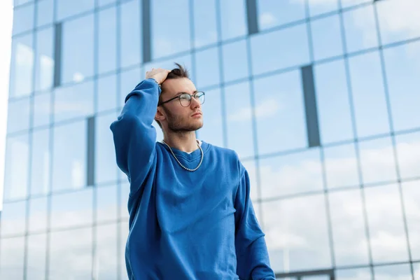 Handsome business man model hipster with hairstyle in fashion blue sweatshirt walks in the city near a modern glasses building