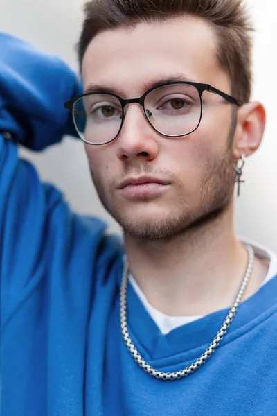 Fashion portrait of a handsome fashionable guy with stylish glasses and a cross earring in a blue sweatshirt on a white background on the street looking at the camera