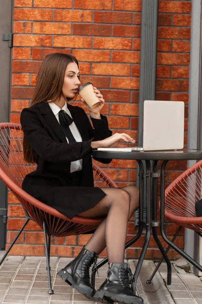 Beautiful professional business woman in fashion black clothes with a skirt and sexy legs with shoes is sitting and working on her laptop, drinking coffee by the brick wall. Trendy female hr manager