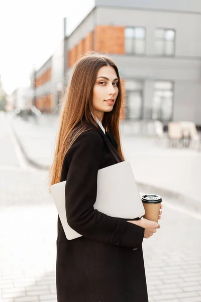 Pretty fashion business woman in black clothes with laptop and coffee to go walks on the street. Female hr manager on the street