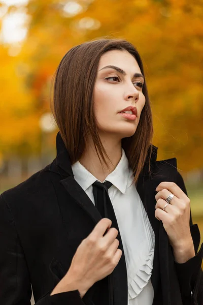 Fresh autumn natural portrait of a beautiful young business woman in a fashion black coat, shirt and tie walking in a yellow autumn park