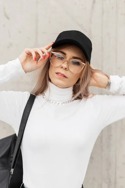 グレーのコンクリート壁の近くにレザーバッグスタンド付きホワイトセーターでメガネとブラックキャップとトレンディなファッション女性 — ストック写真