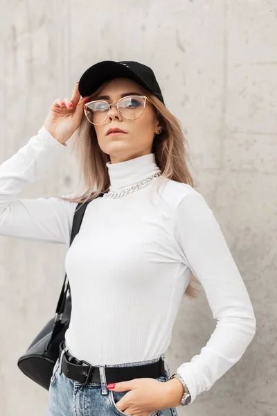 ファッショナブルなブラックキャップとバックパック付きの白いセーターを身に着けているメガネを持つスタイリッシュな美しい若い女性は 市内の灰色のコンクリート壁の近くに立つ — ストック写真