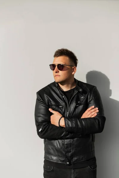 Fashion young cool brutal man in black stylish clothes with cool sunglasses, black leather jacket stands near a gray wall in the street