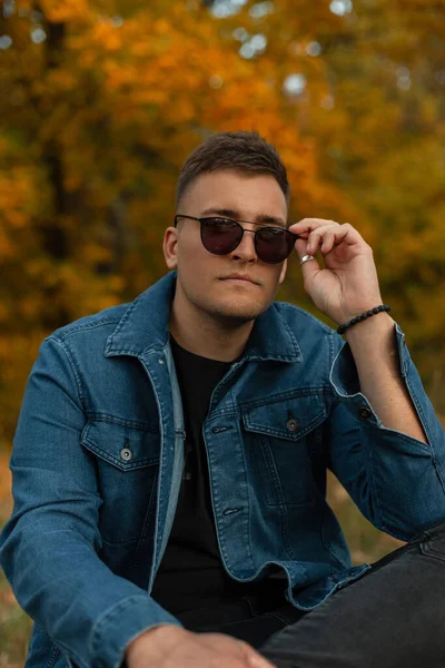 fashionable handsome young man model in trendy denim clothes sits in an autumn park and wearing vintage sunglasses. Fall male stylish look clothes