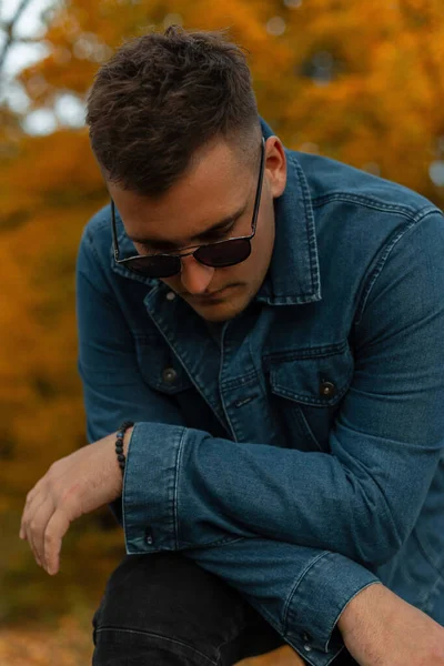 Fashion male portrait of handsome cool man with hairstyle in jeans clothes with sunglasses poses in autumn colorful park
