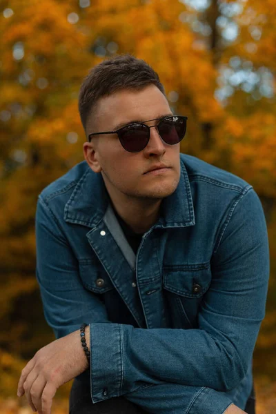 Fashionable handsome man model with hairstyle in a denim shirt with black sunglasses in nature against a background of yellow autumn leaves in the park