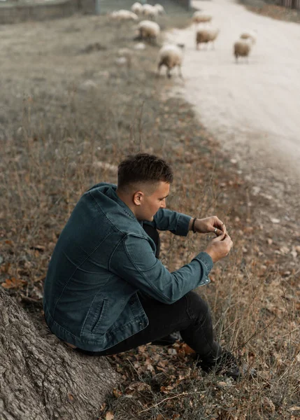 Fashionable handsome hipster country man in a stylish blue jeans jacket sits on the grass near the road and a flock of sheep in the countryside