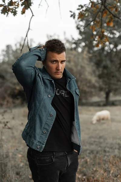 Fashion young country hipster man with hairstyle in stylish denim shirt and black jeans stands outdoors in countryside