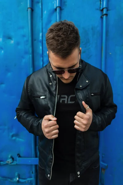 Handsome fashion young man with hairstyle and sunglasses in trendy black leather jacket and t-shirt stands and poses near a blue metal background