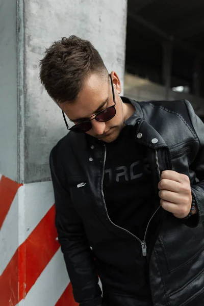 Stylish street man model with hair with black sunglasses wearing black clothes with a black leather jacket in the city. Urban male style look clothes